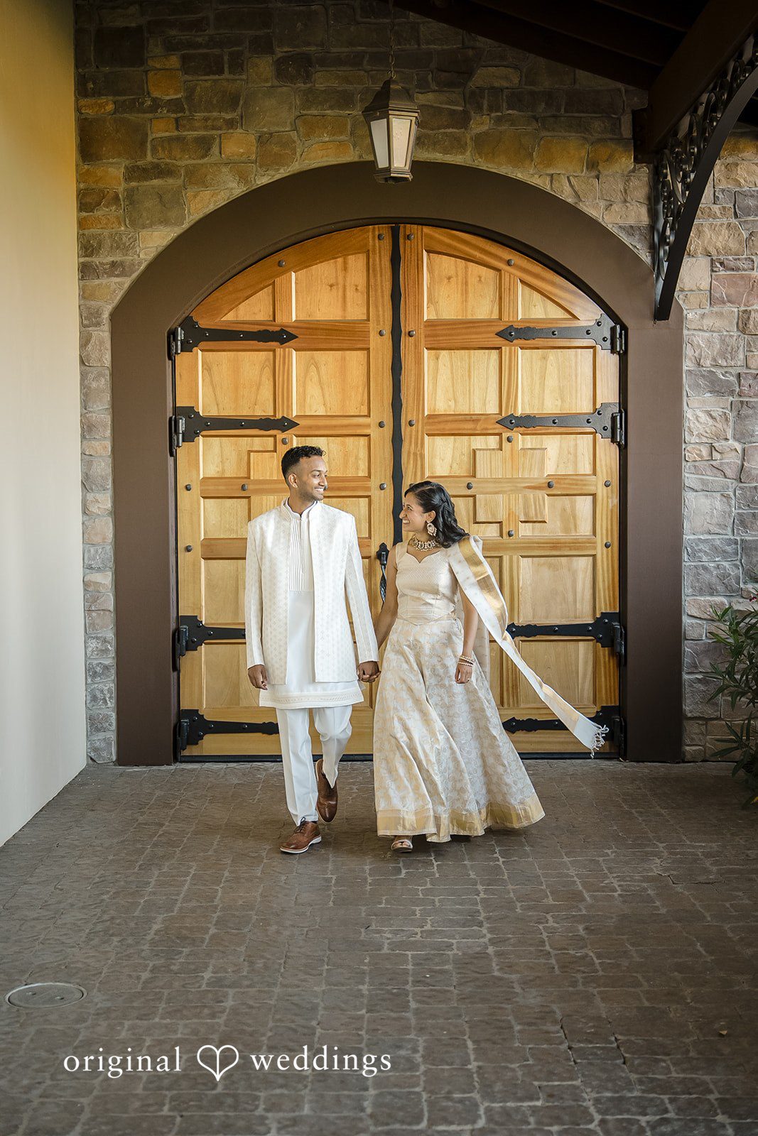 Kathir + Nandita A bride and groom holding hands while posing near a rustic stone building.