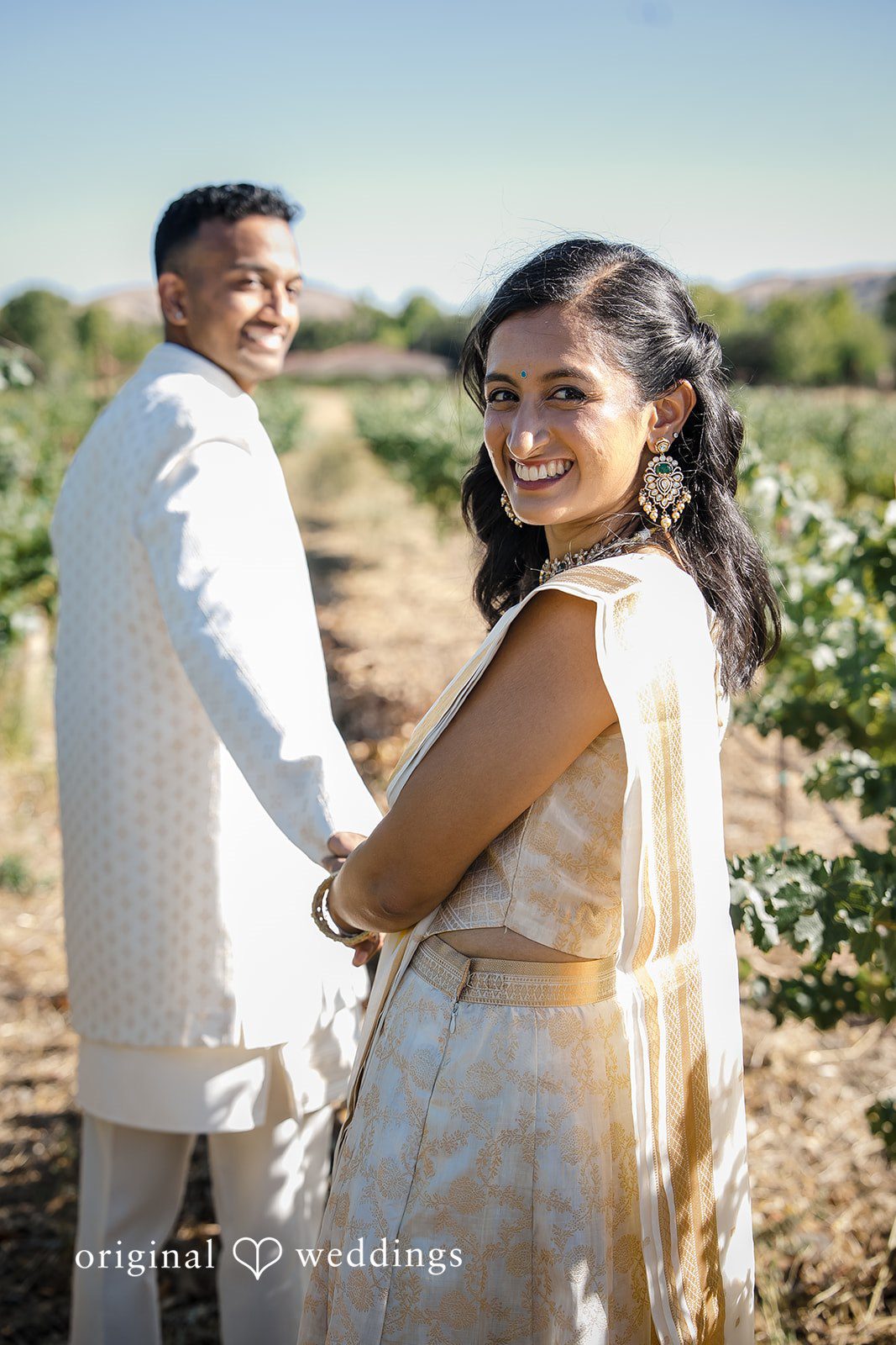 Kathir + Nandita A bride and groom smiling while standing close together in a vineyard setting.