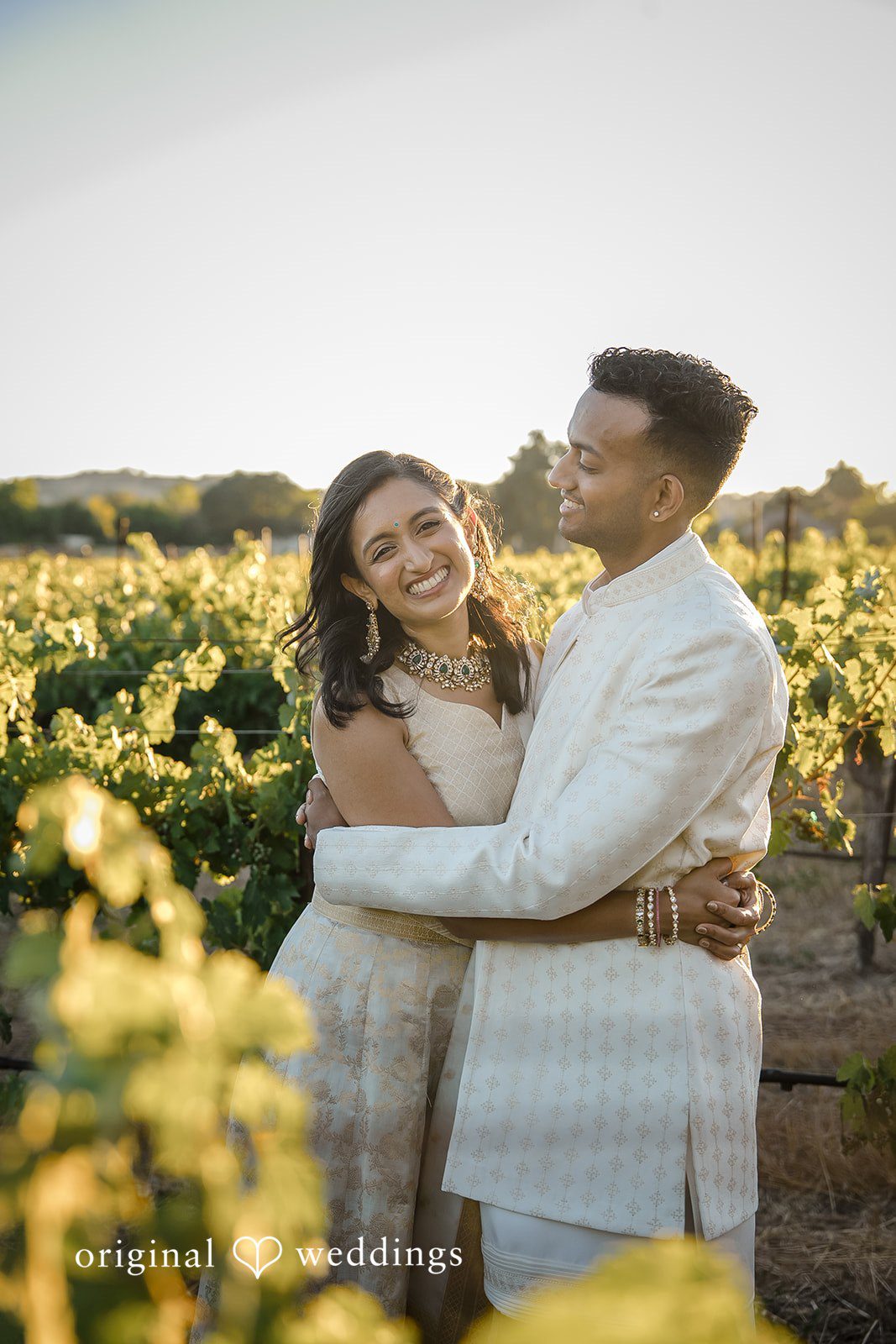 Kathir + Nandita A bride and groom posing affectionately in front of sunlit vineyard rows.