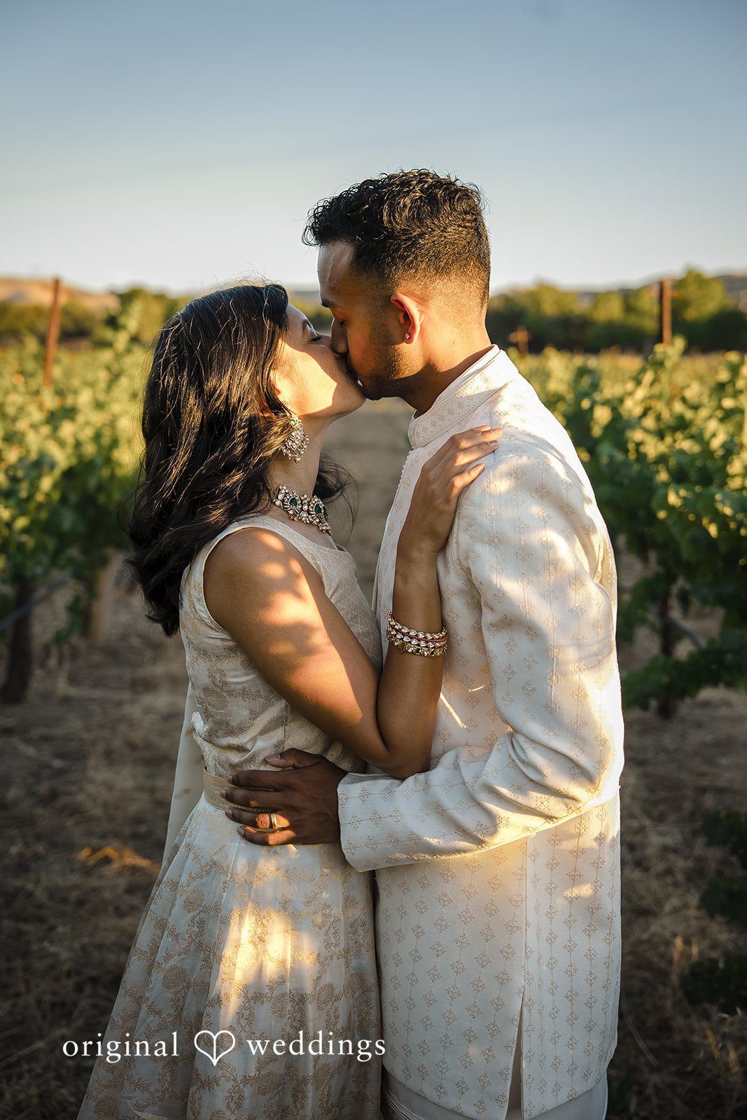 Kathir + Nandita A bride and groom sharing a warm embrace in a sunlit outdoor landscape.