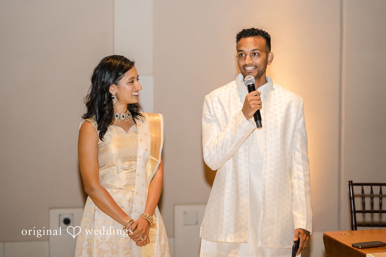 Kathir + Nandita A man speaking into a microphone next to a woman during a reception event.