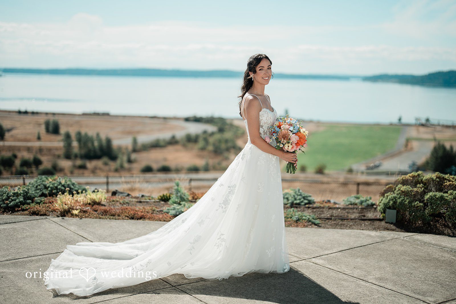 Katie + Gerald A bride in a long white gown posing on a grassy hill overlooking the water.