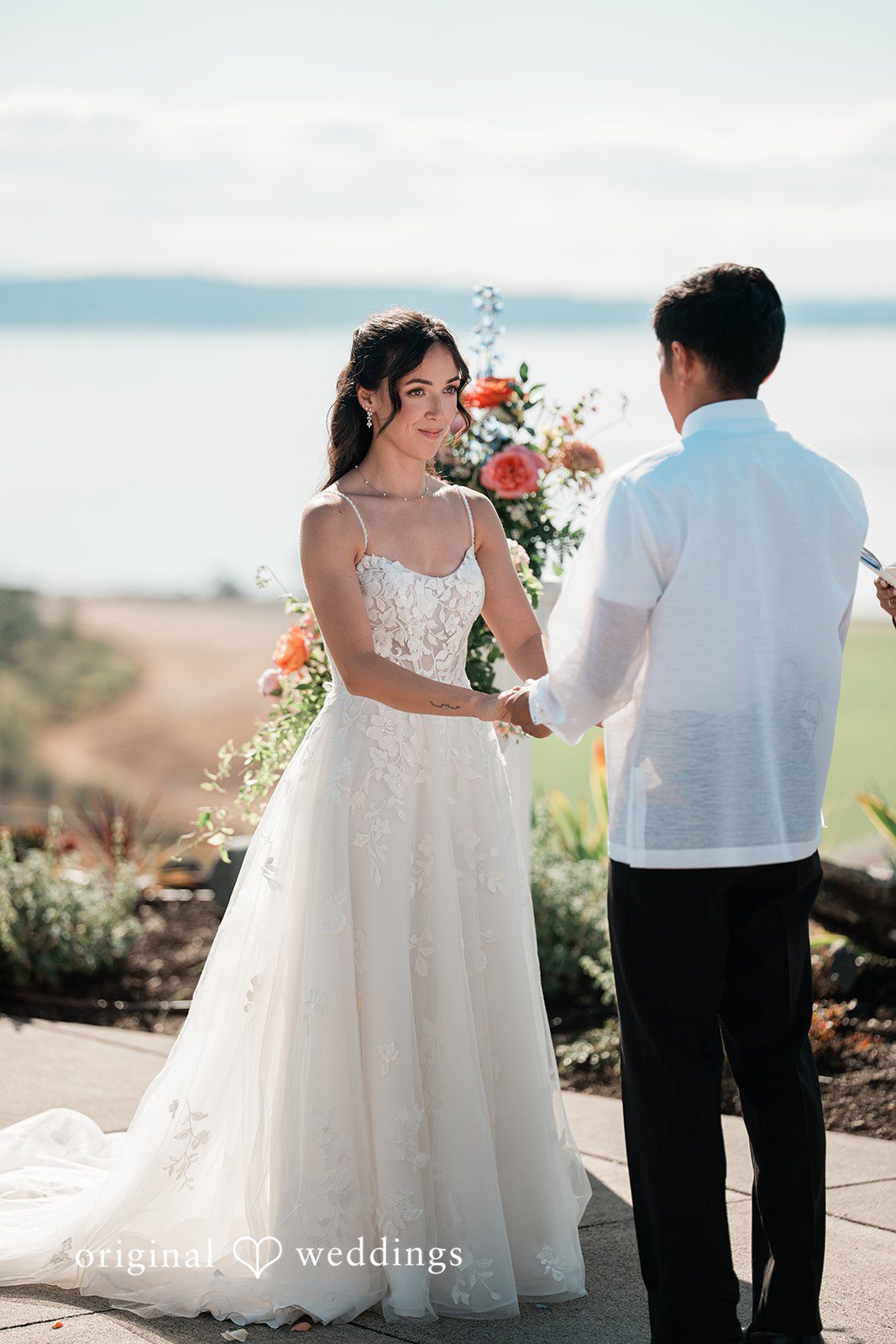 Katie + Gerald A bride and groom holding hands and smiling at each other by the coast.