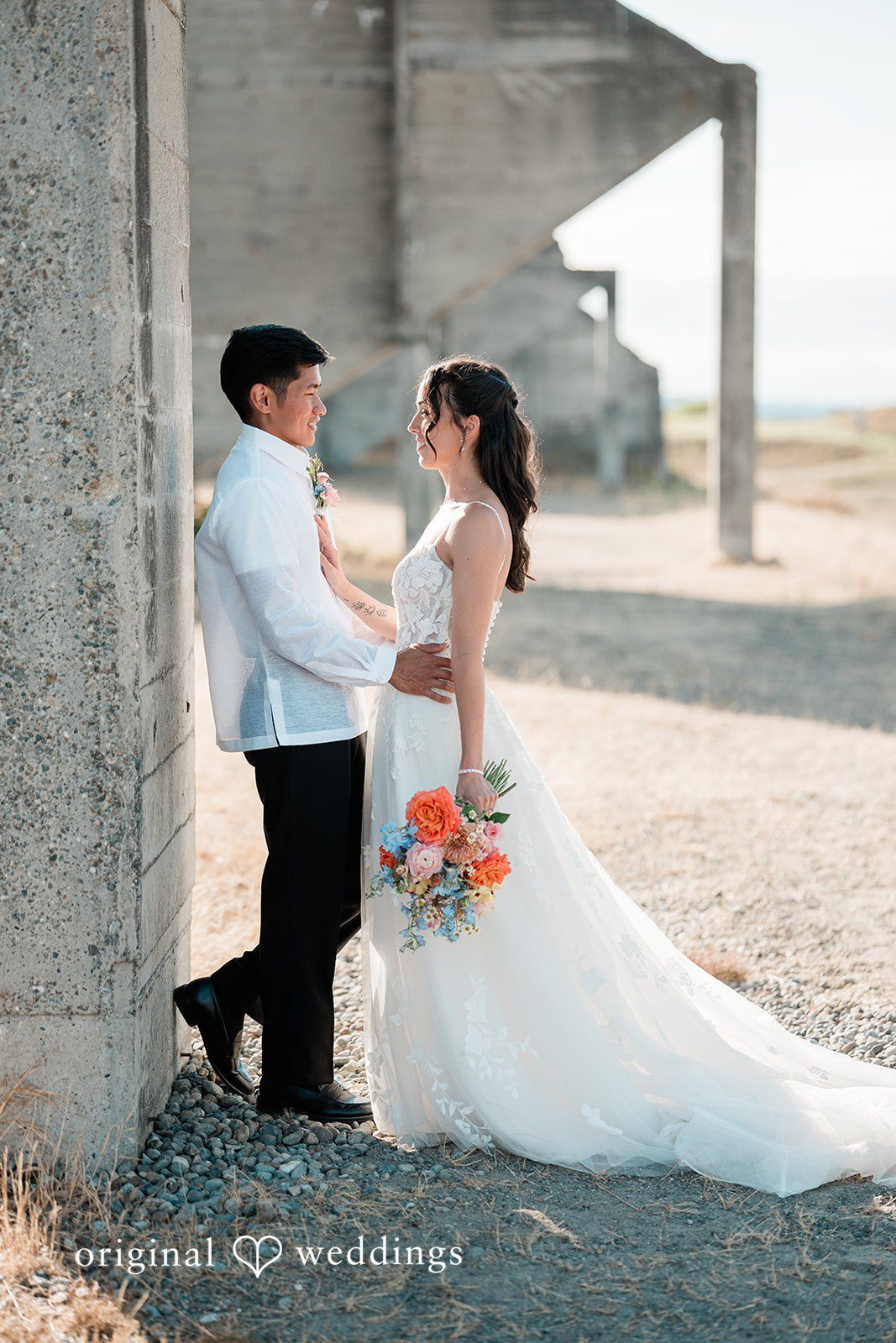 Katie + Gerald A bride and groom walking hand-in-hand next to an open concrete building.