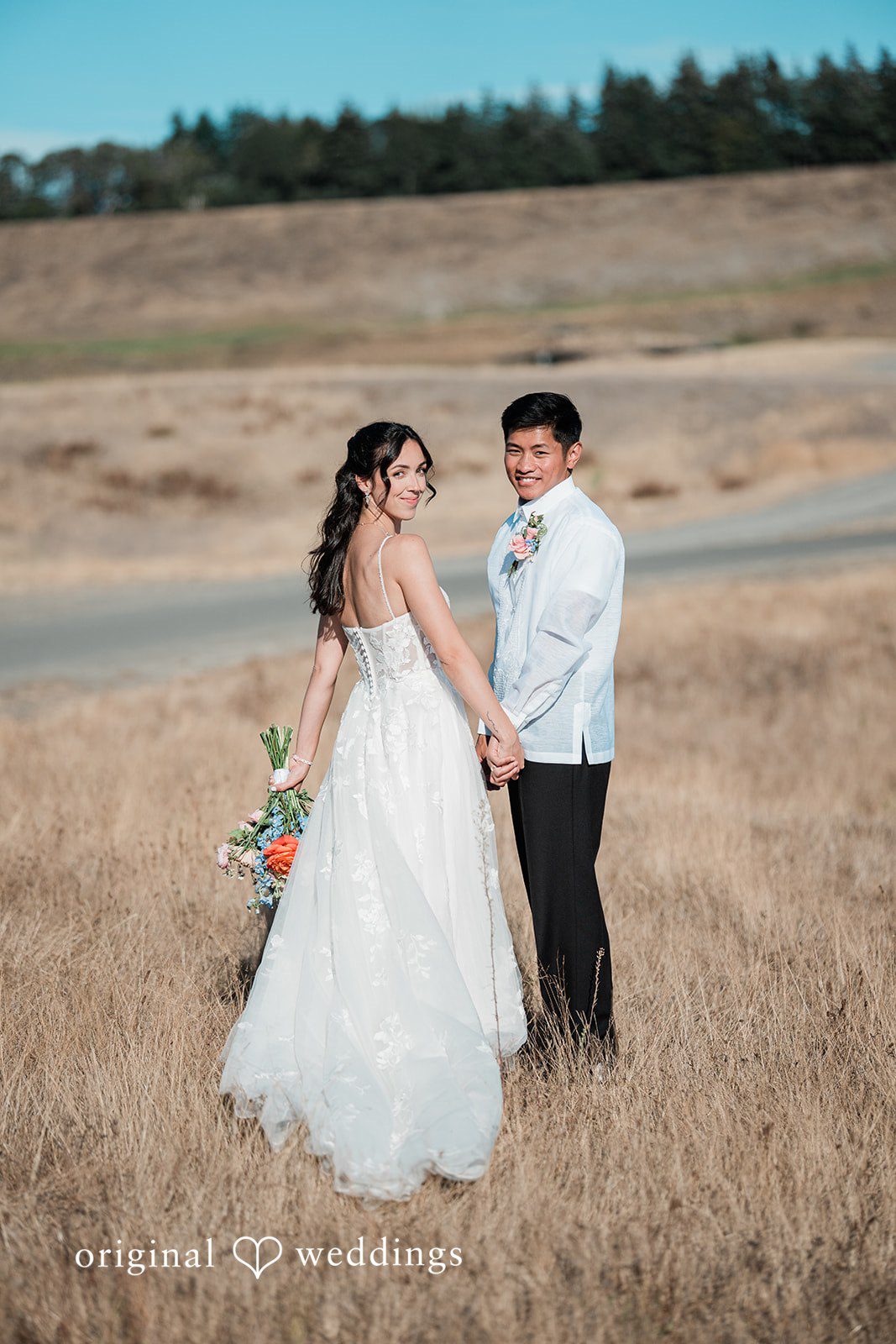 Katie + Gerald A bride and groom walking hand-in-hand through an open grassy field.
