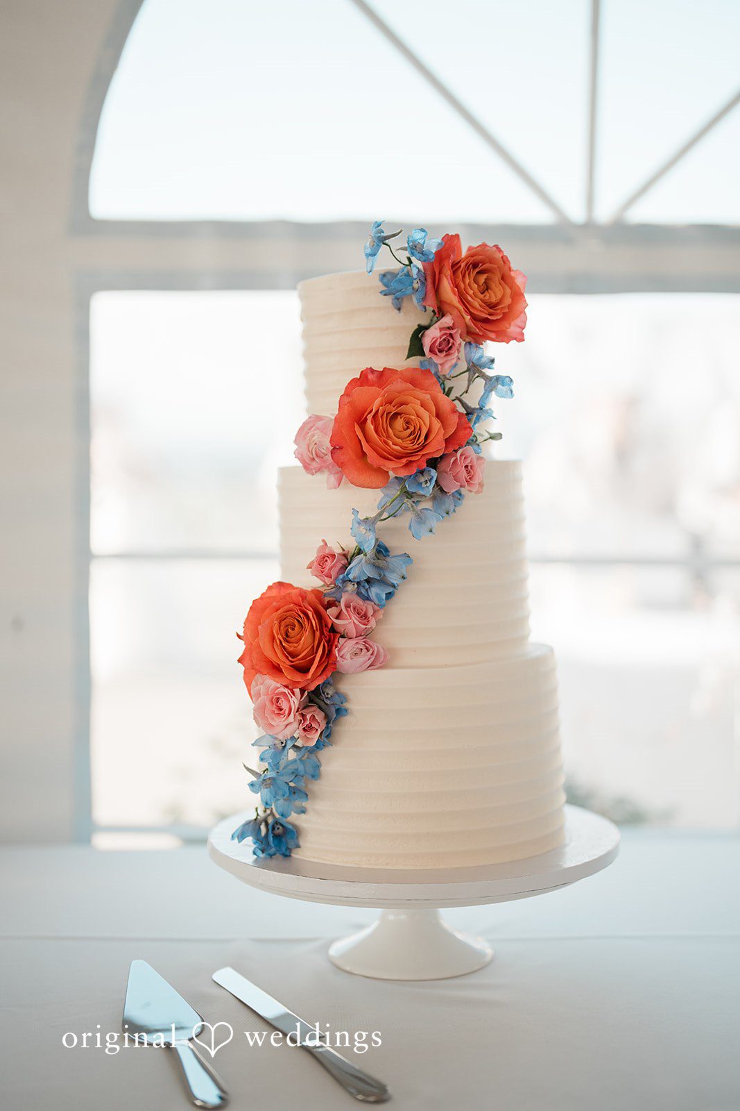 Katie + Gerald A multi-tiered white wedding cake decorated with a cascade of blue and orange flowers.