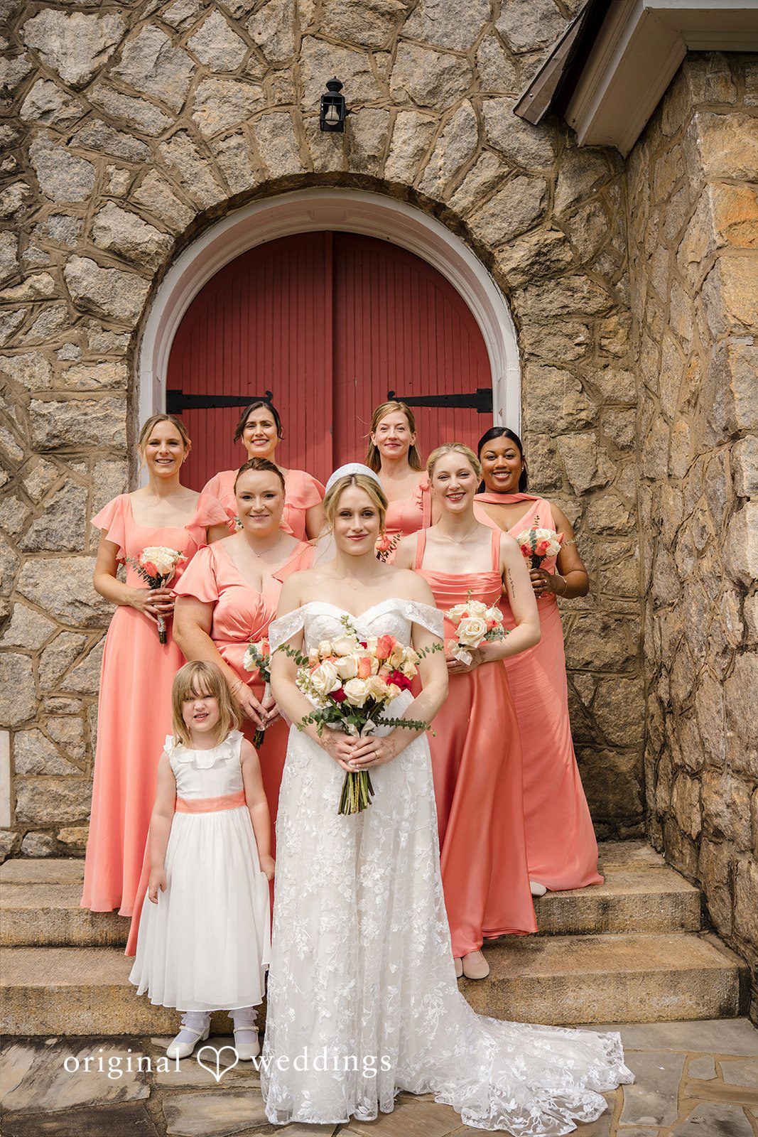 Julie + Joshua A bride and her bridesmaids in matching pink dresses posing together in front of a stone wall.