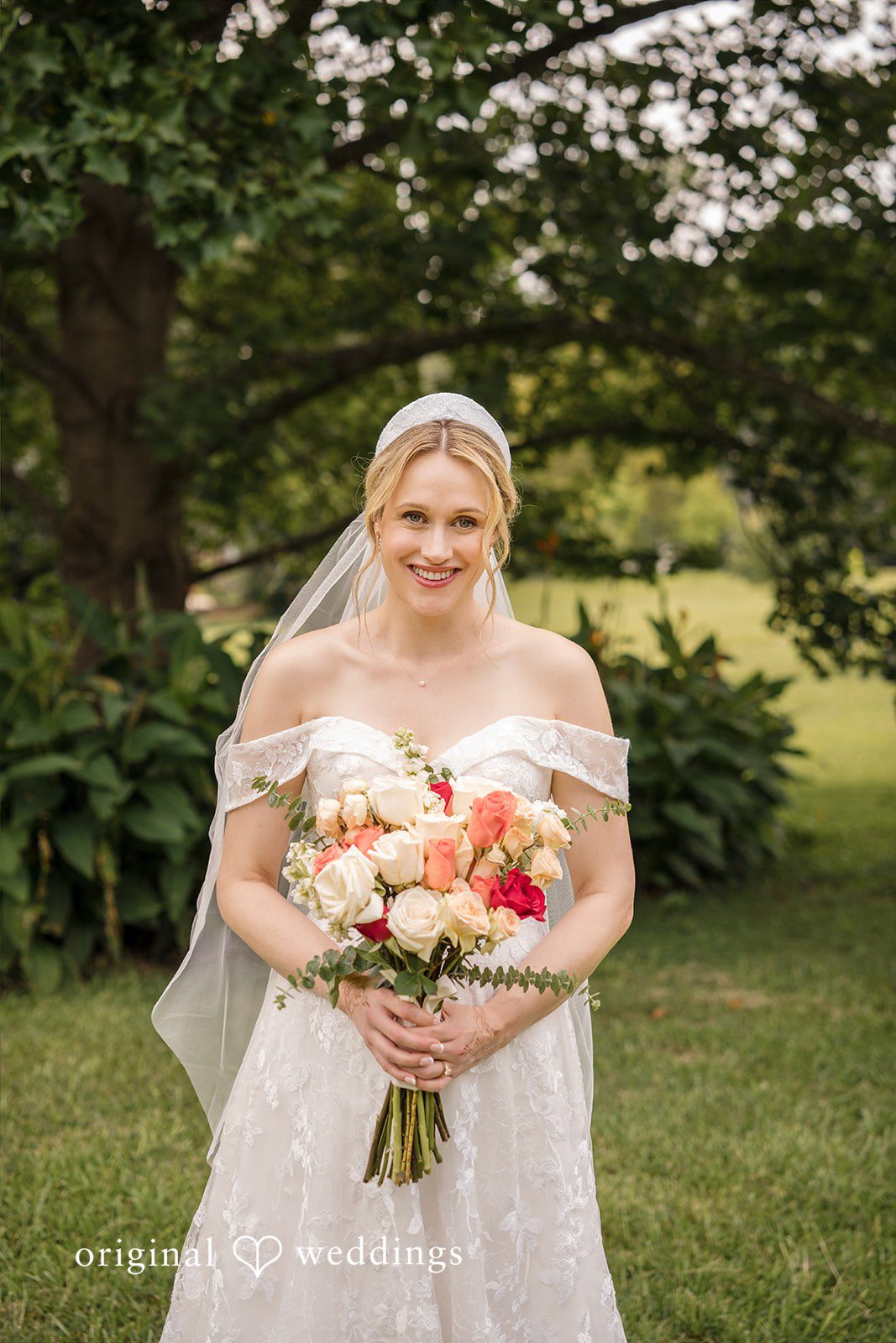 Julie + Joshua A smiling bride in a lace gown holding a bouquet in front of a green background.
