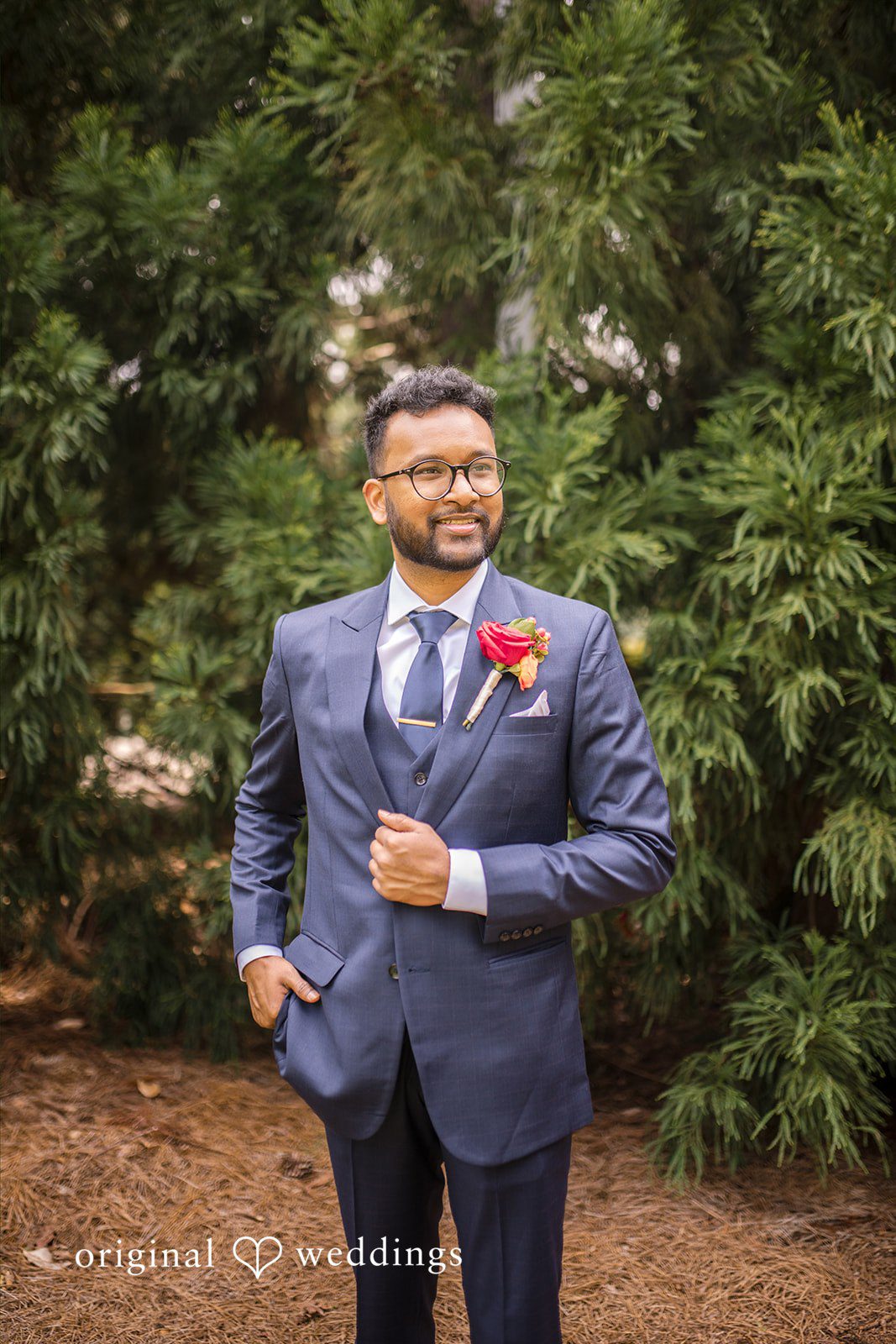 Julie + Joshua A groom in a blue suit smiling while posing against a backdrop of green foliage.