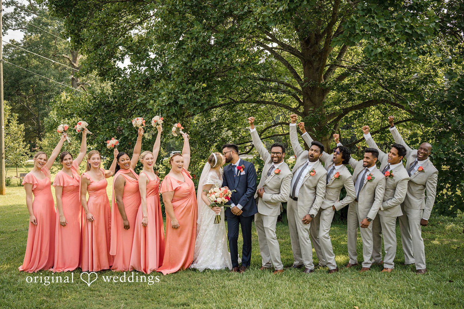Julie + Joshua A large wedding party posing together on a grassy field in front of a forest line.