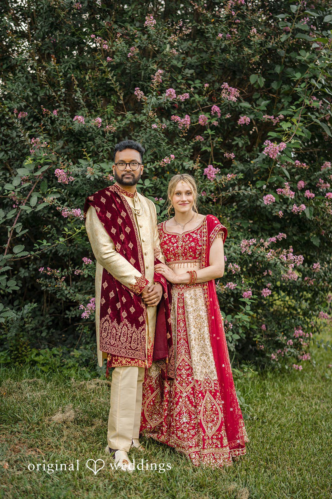 Julie + Joshua A bride and groom posing side-by-side in matching traditional red and cream outfits.