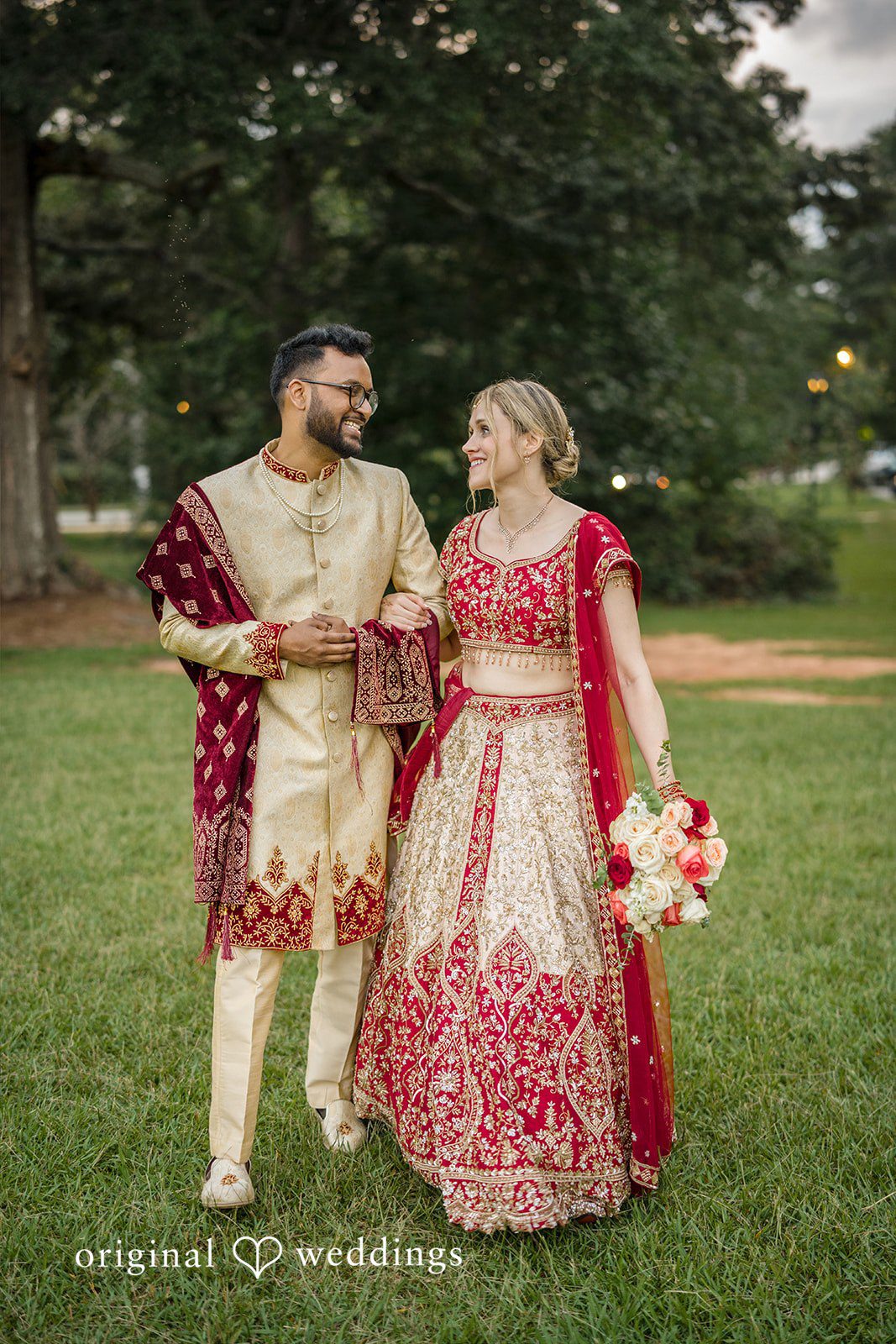 Julie + Joshua A bride and groom in traditional red clothing walking together through a park.