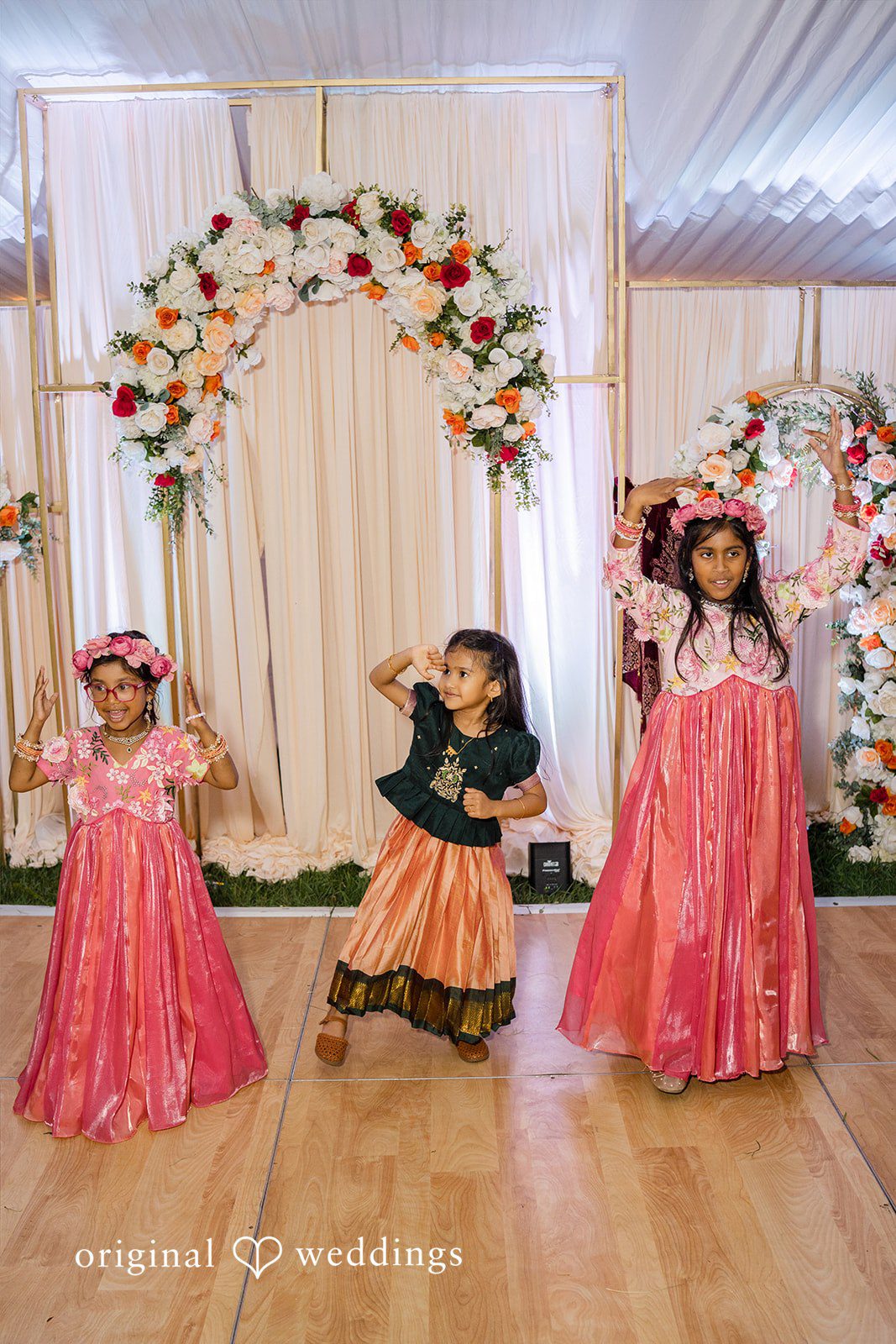 Julie + Joshua Three children in traditional pink outfits standing together at a festive event.