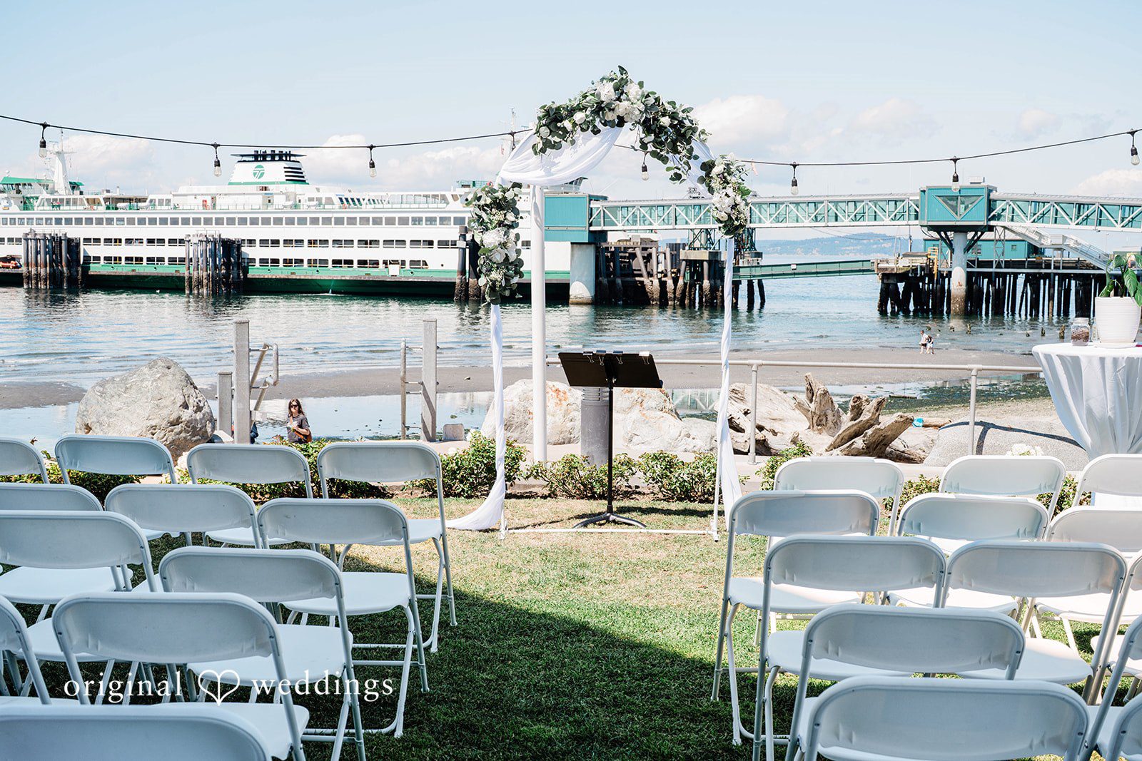 Naomi + Darrell Waterfront wedding ceremony setup with white chairs and a floral arch.