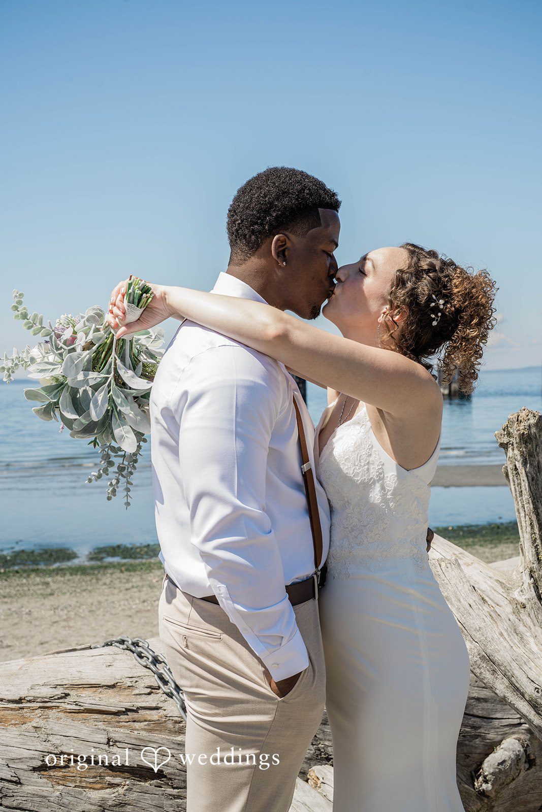 Naomi + Darrell Groom embracing the bride from behind on a sandy beach.