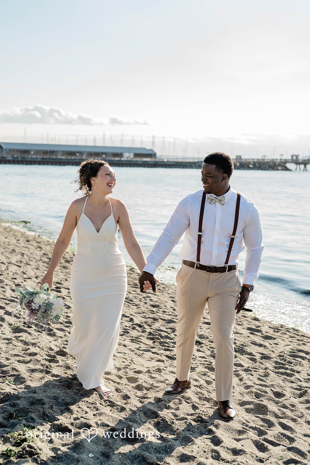 Naomi + Darrell Bride and groom walking hand-in-hand along a coastal walkway.