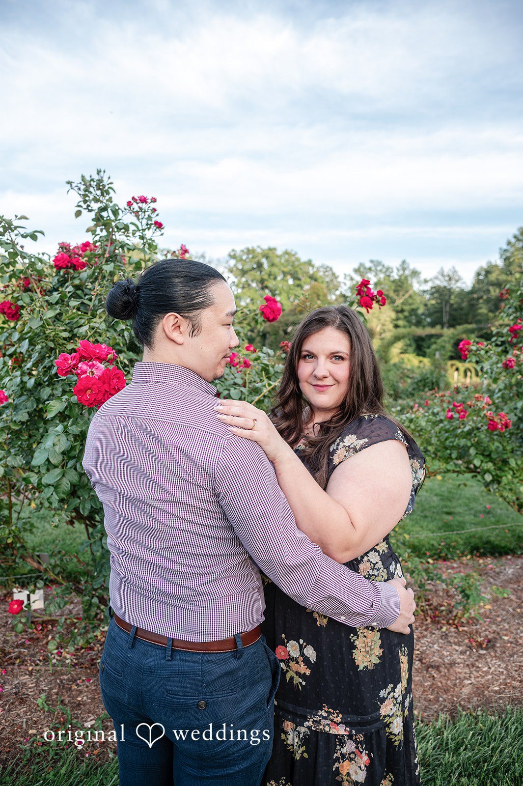 Gabrielle + James A man embracing a woman from behind as they pose together in a rose garden.