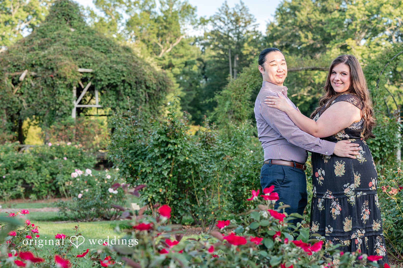 Gabrielle + James A couple holding each other and smiling while standing among lush green bushes and red flowers.