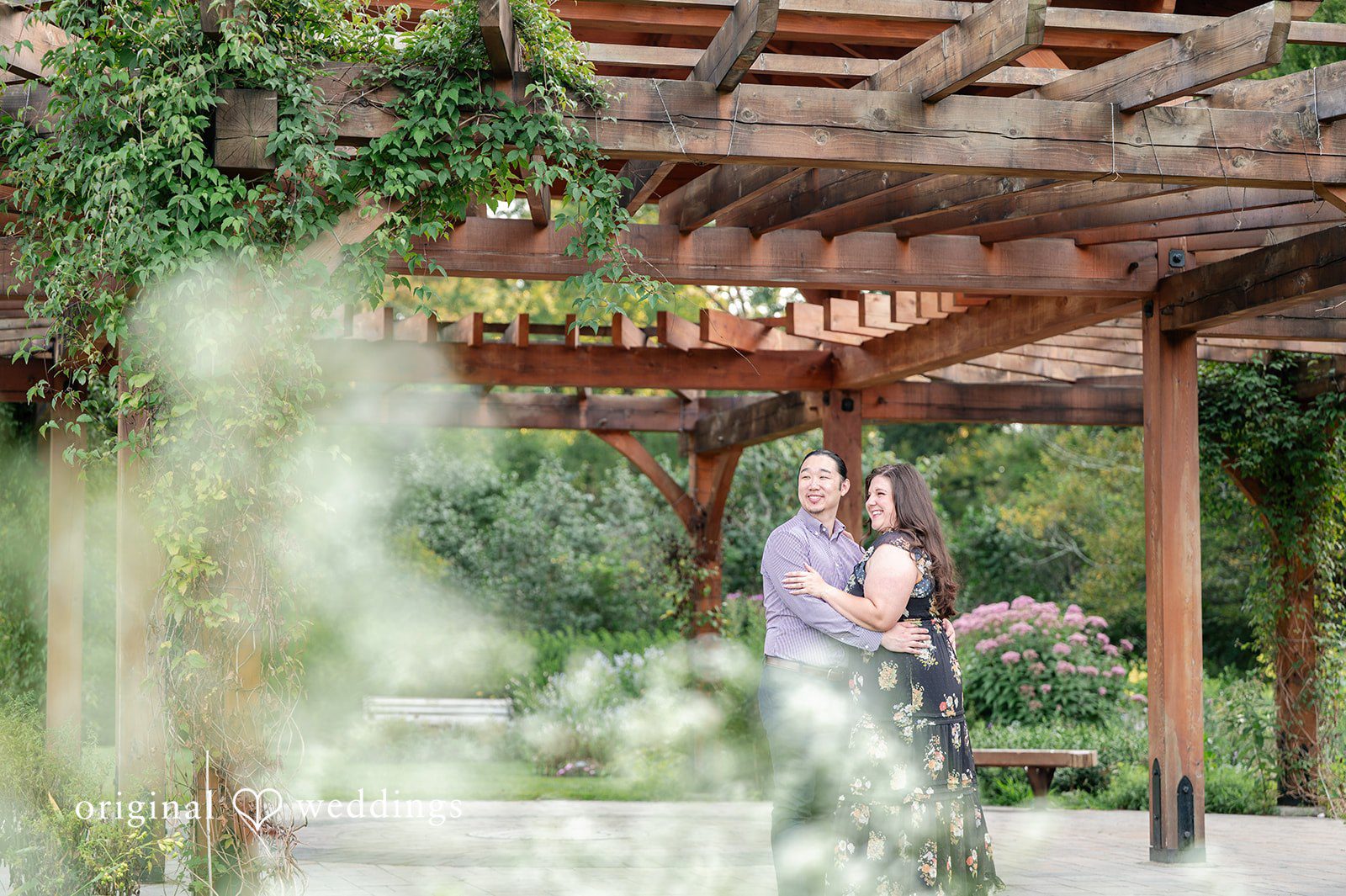 Gabrielle + James A couple posing under a long wooden pergola covered in climbing green vines.