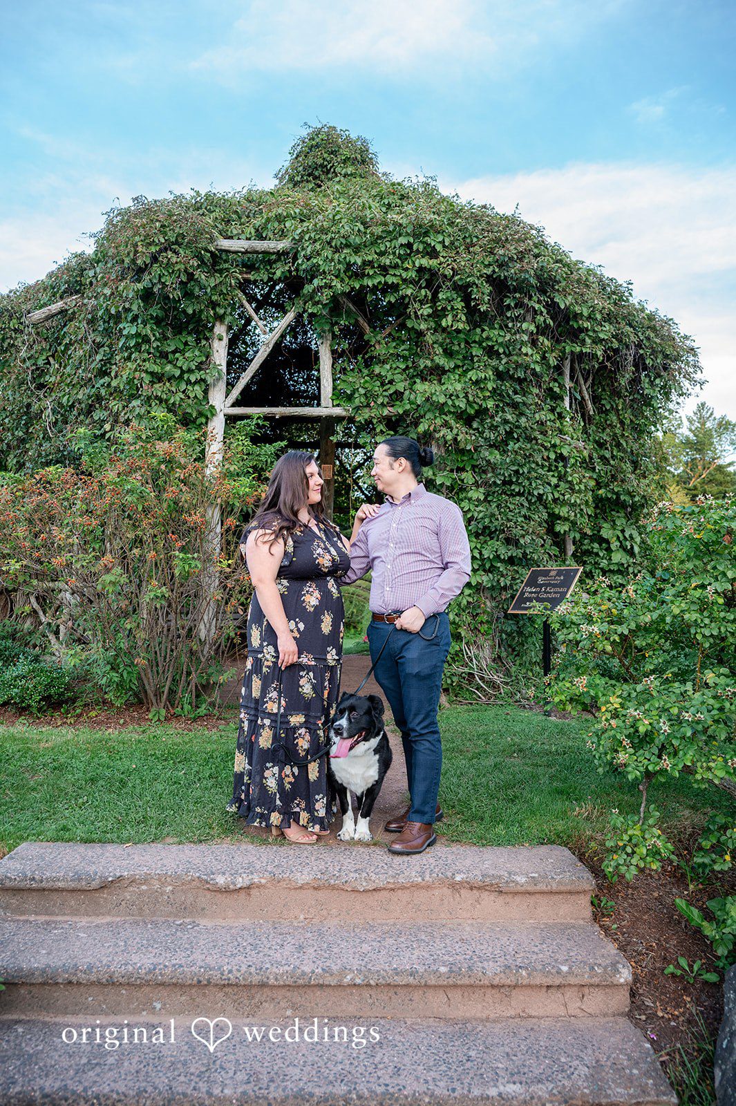 Gabrielle + James A couple standing together on steps in front of a massive wall of green foliage.