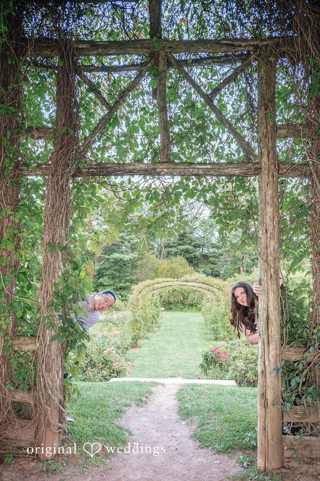 Gabrielle + James A couple posing in the distance framed underneath a large wooden garden trellis archway
