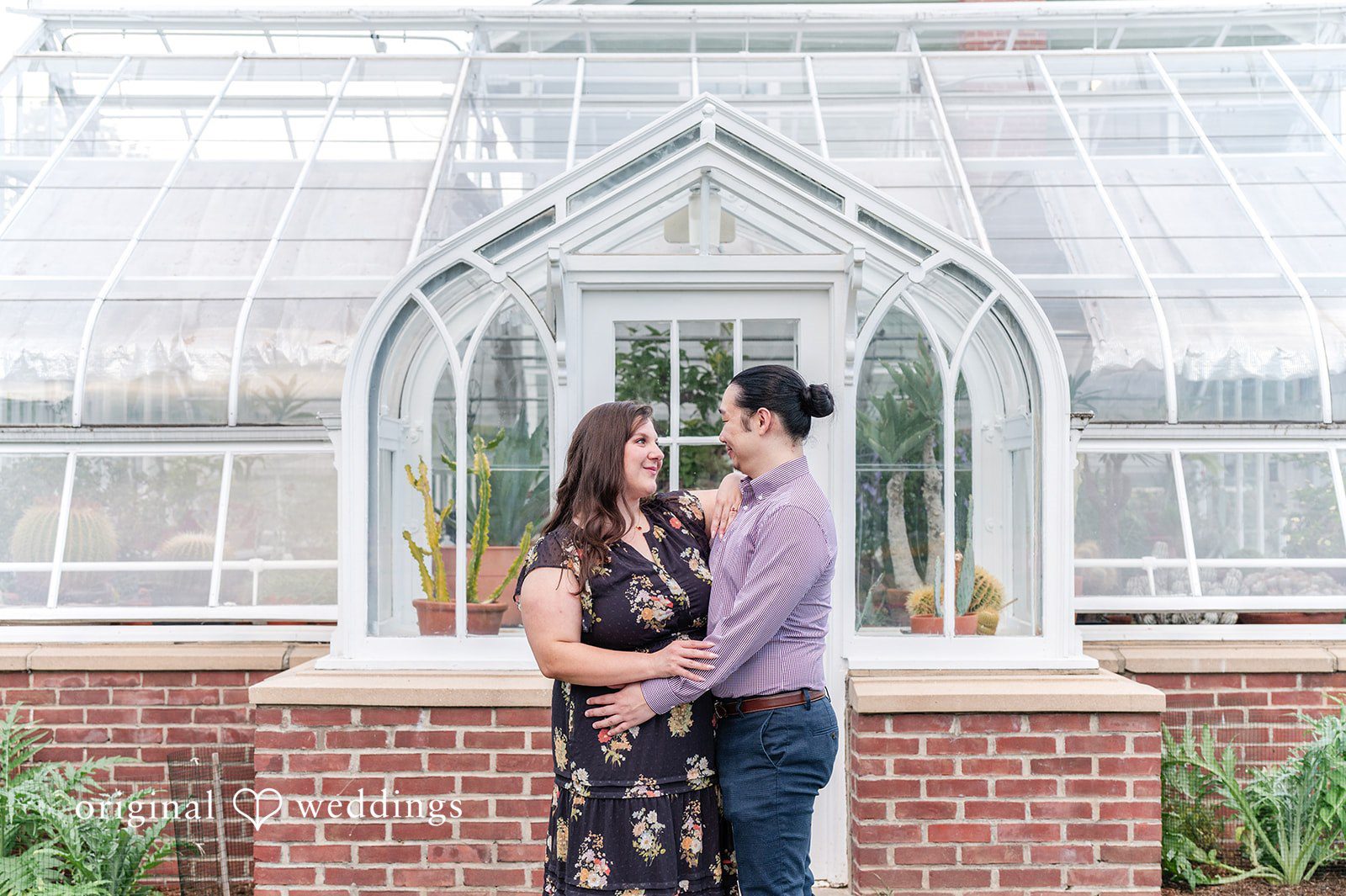 Gabrielle + James A couple posing closely in front of the entrance of a white glass greenhouse structure.
