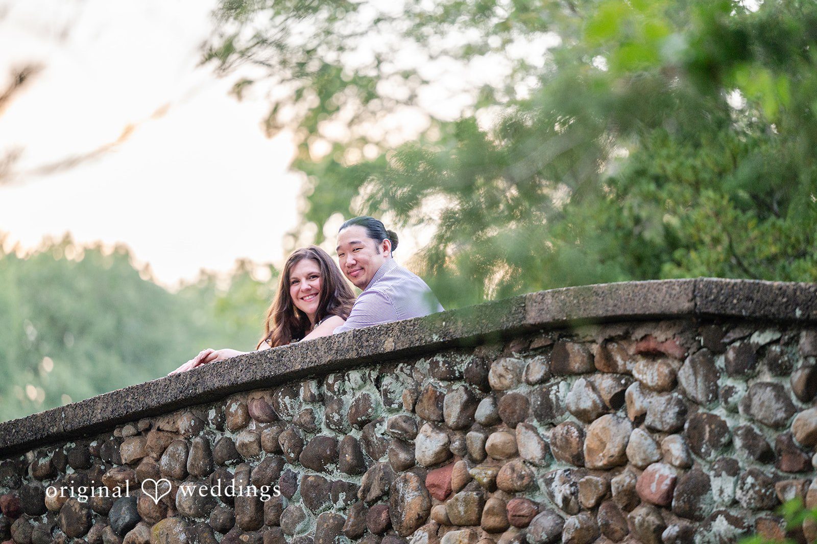 Gabrielle + James A couple leaning against a rustic stone bridge wall surrounded by greenery.
