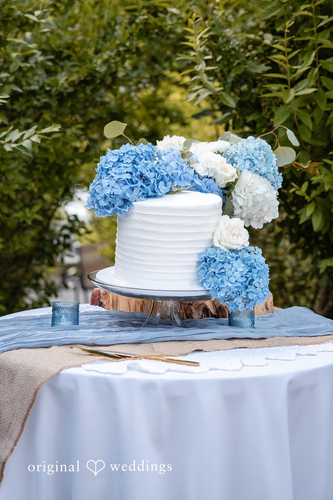 July 17, 2026 – Alissa & Duncan – (ID-5192) – REVISED 9.1 A white wedding cake decorated with vibrant blue hydrangeas on a fabric-covered table.