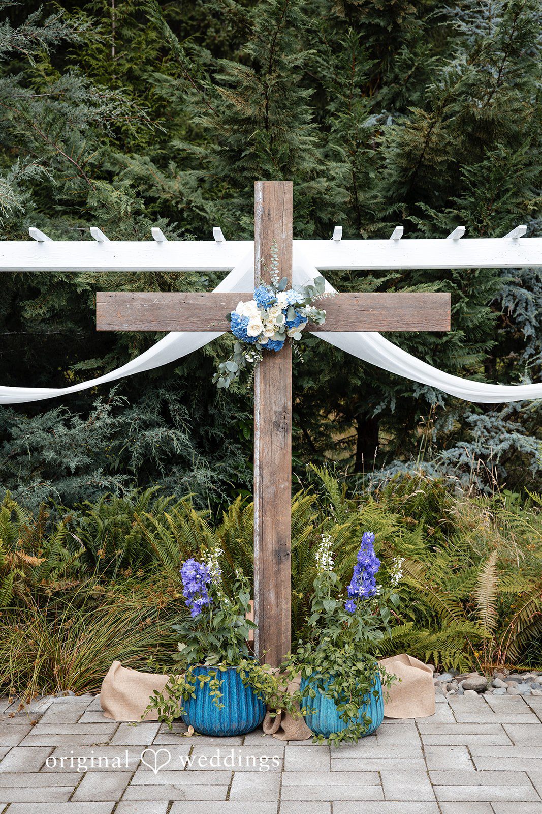 Alissa + Duncan A large wooden cross decorated with white fabric and flowers at a ceremony site.