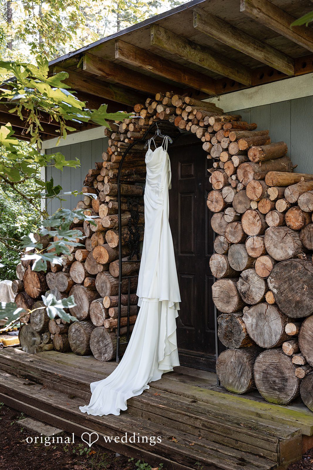 Alissa + Duncan A white wedding dress hanging against a rustic backdrop of stacked firewood logs.