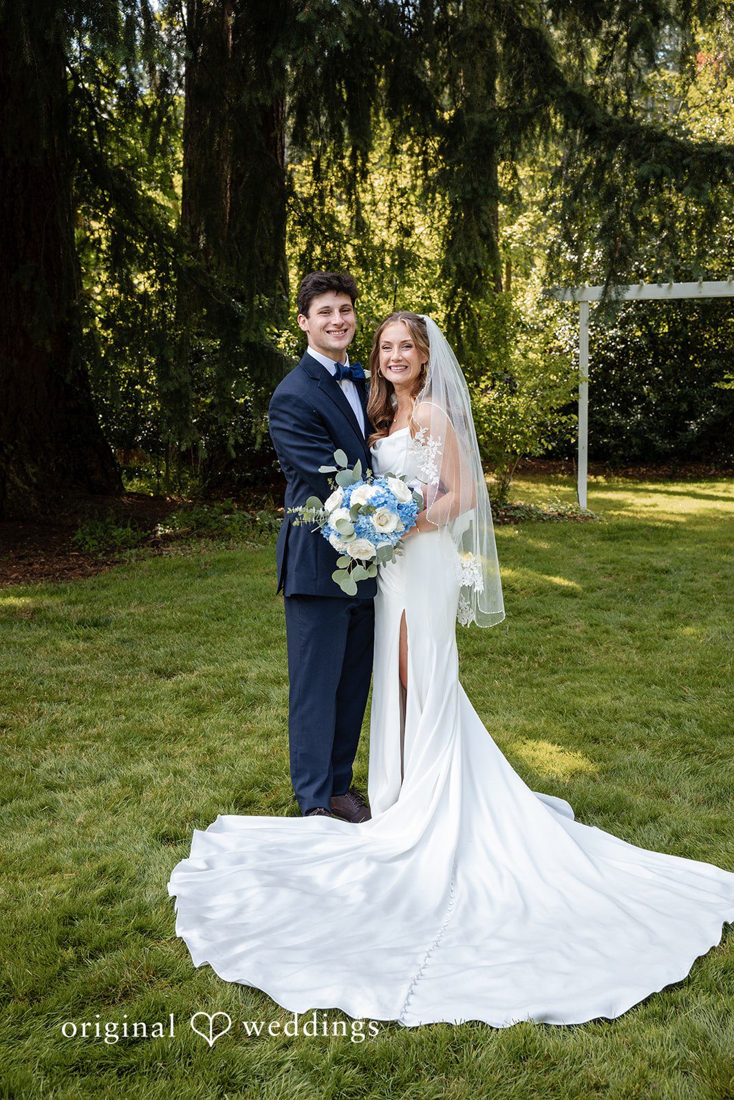 Alissa + Duncan A bride and groom posing together as her long white dress train spreads across the lawn.