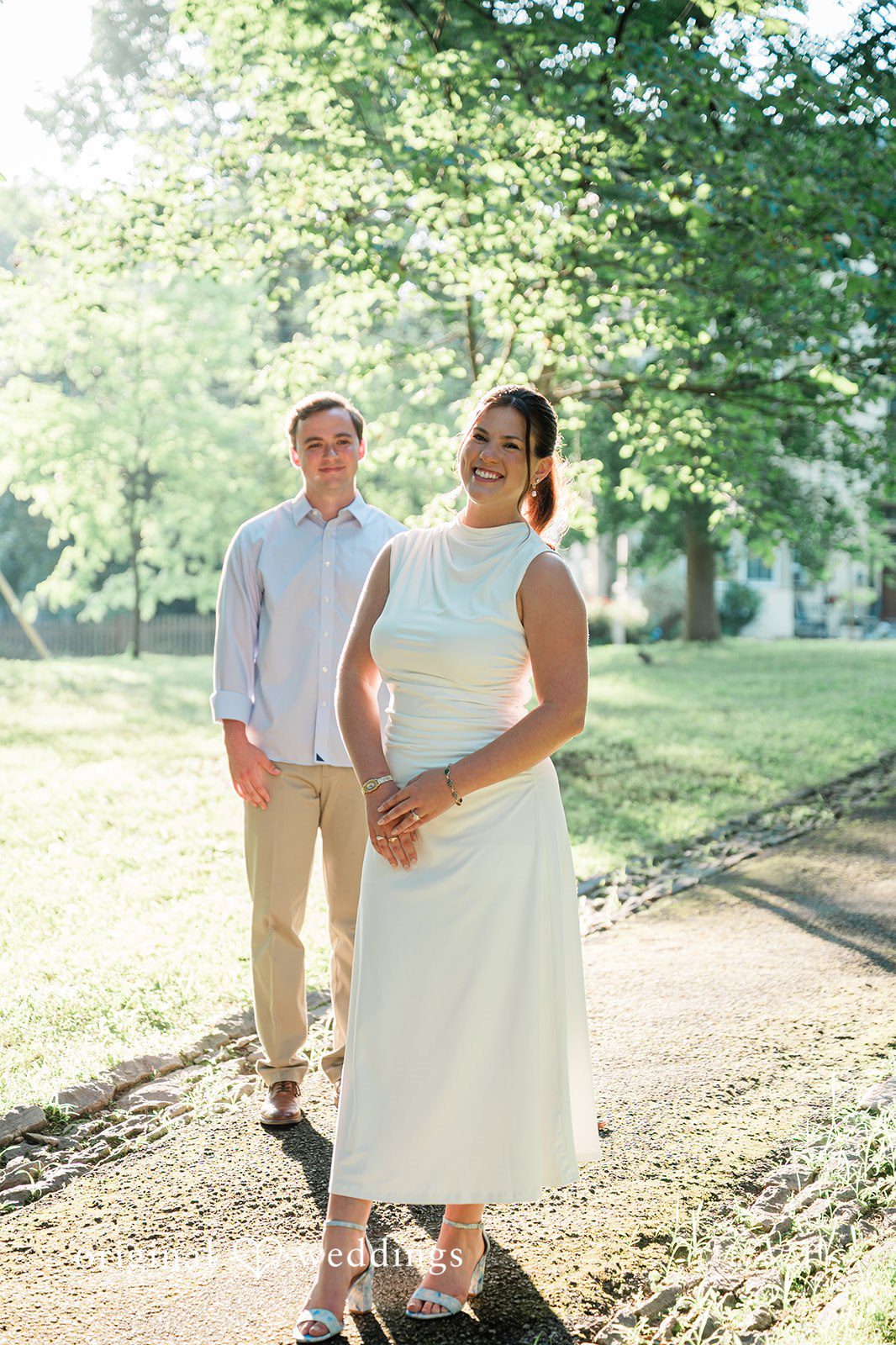 A couple standing together near a wooded path, smiling happily.