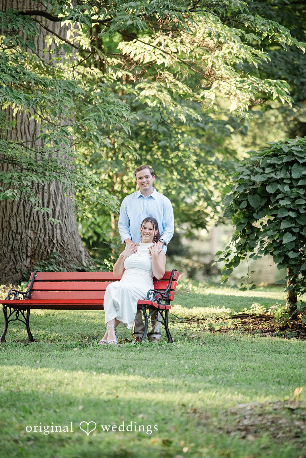 A couple sitting together on a red park bench surrounded by green foliage.