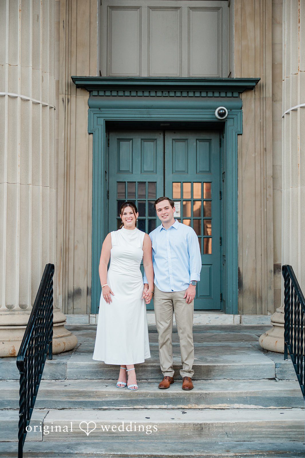 A couple standing side-by-side in front of a large, classic green entrance door.