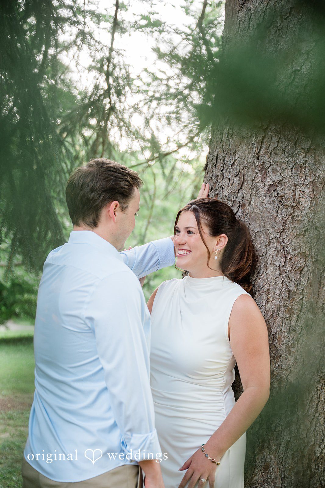 A man and a woman smiling at each other while leaning against a large tree trunk.