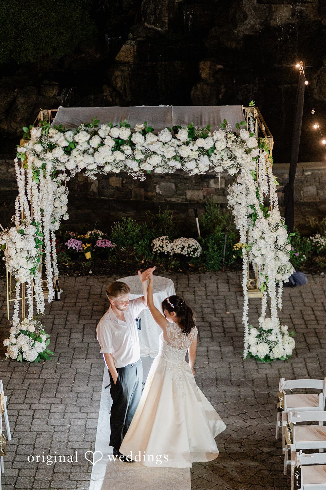 Julia + Camden A bride and groom hold hands and dance on a white aisle runner beneath a large floral arch decorated with white flowers.