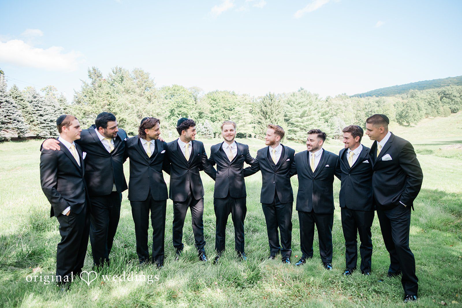 Julia + Camden A group of groomsmen in black suits standing together on a grassy hill.