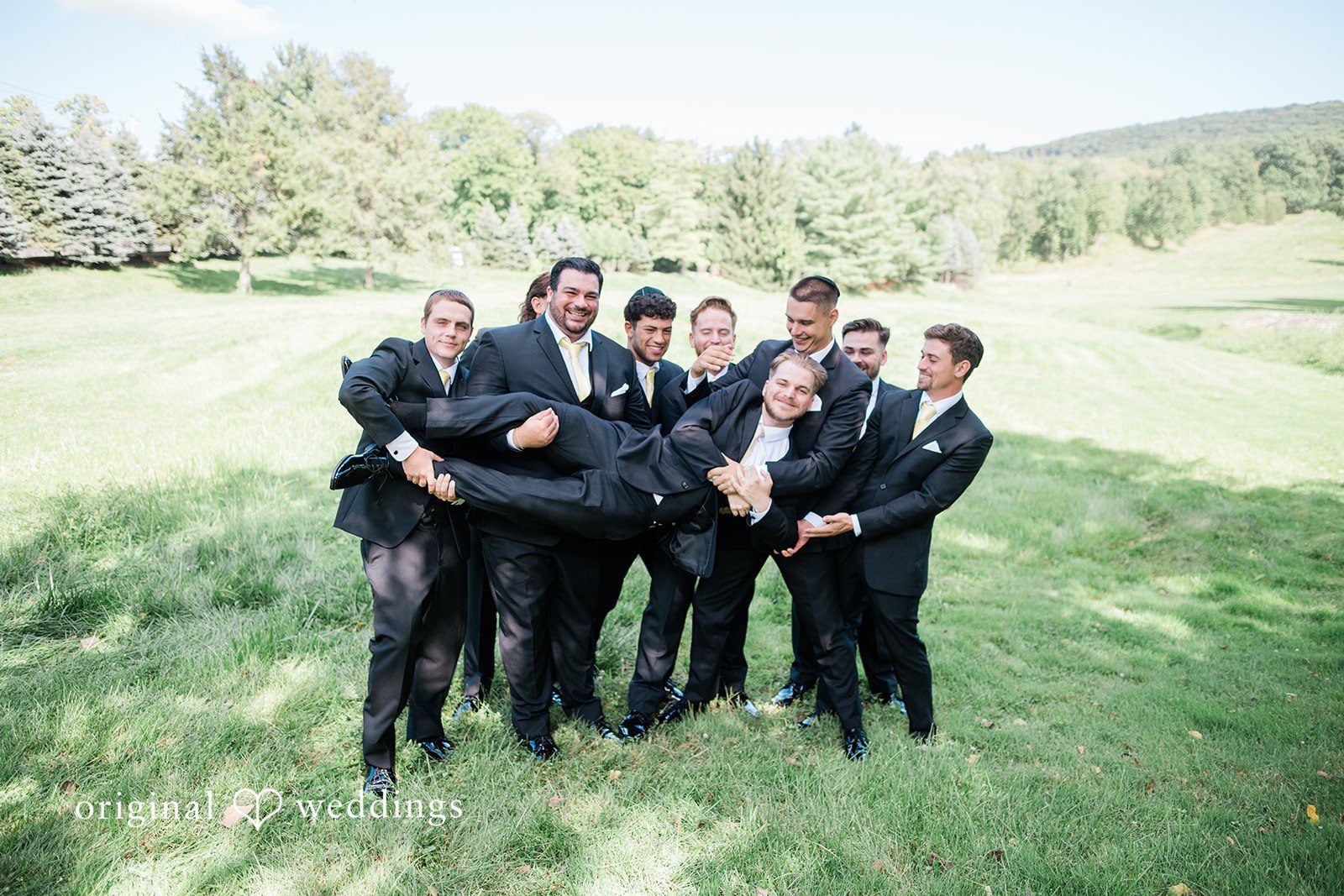 Julia + Camden A groom being lifted into the air by his groomsmen on a grassy field.