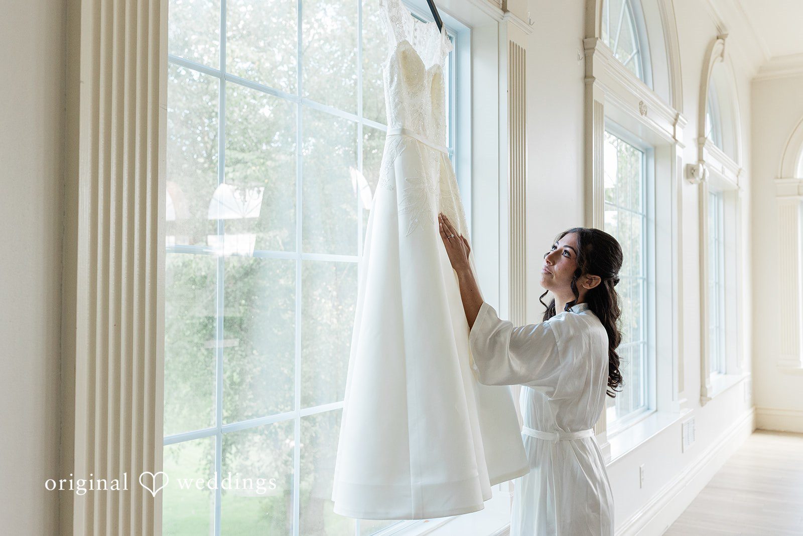 Julia + Camden A bride in a white gown standing next to a large window looking outward.