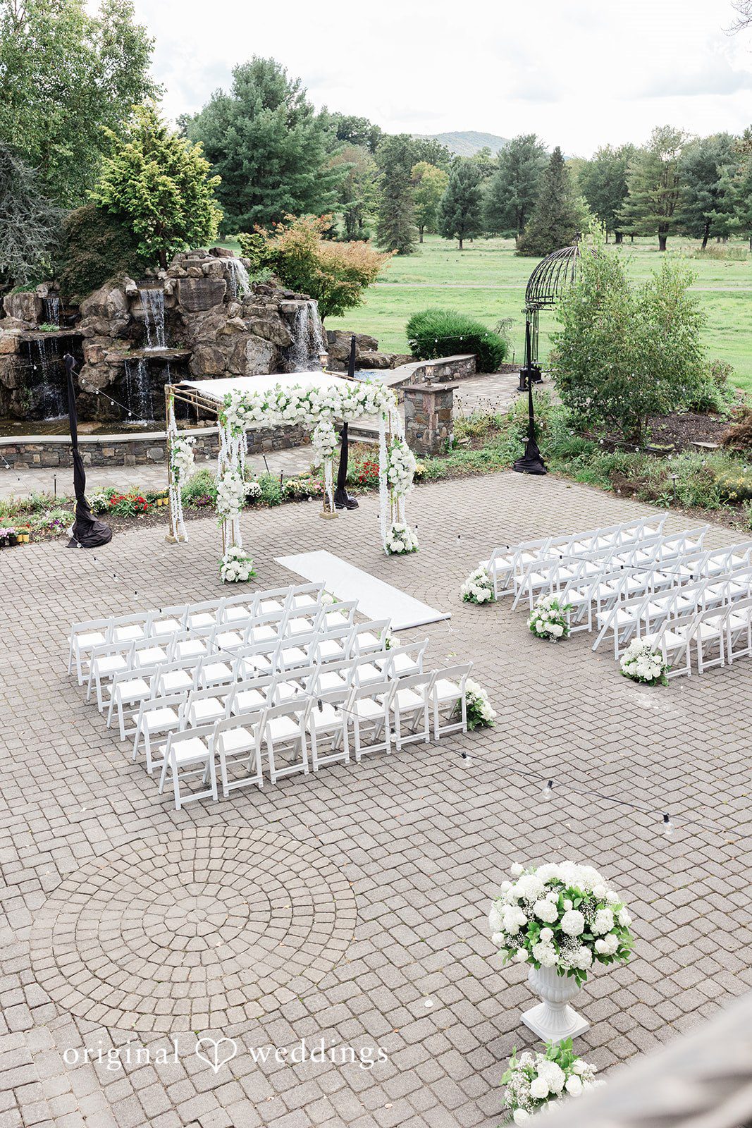 Julia + Camden Rows of white chairs set up for a ceremony on a gravel path next to a stone structure.