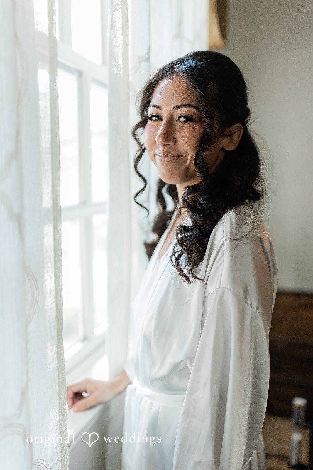 Julia + Camden A bride in a white robe smiling while standing indoors near a window.