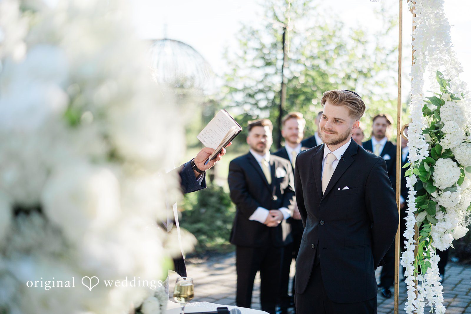 Julia + Camden A groom standing outside surrounded by white floral arrangements and greenery.