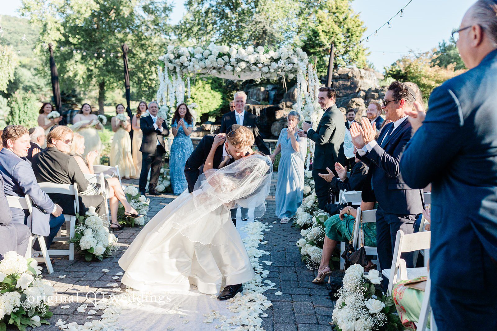 Julia + Camden A bride walks down a white aisle runner between rows of seated guests.