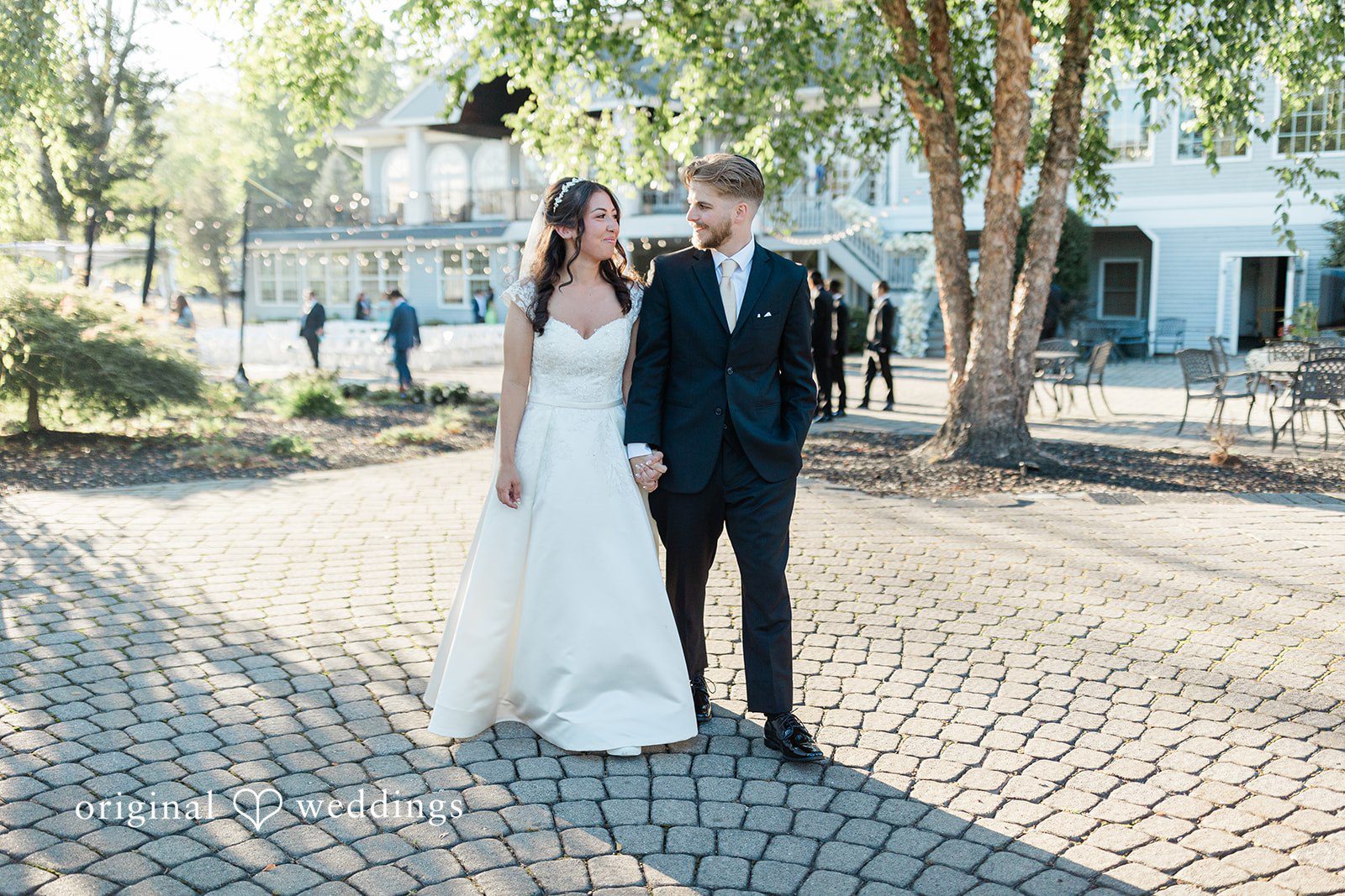Julia + Camden A bride and groom walk hand-in-hand along a paved pathway lined with greenery.