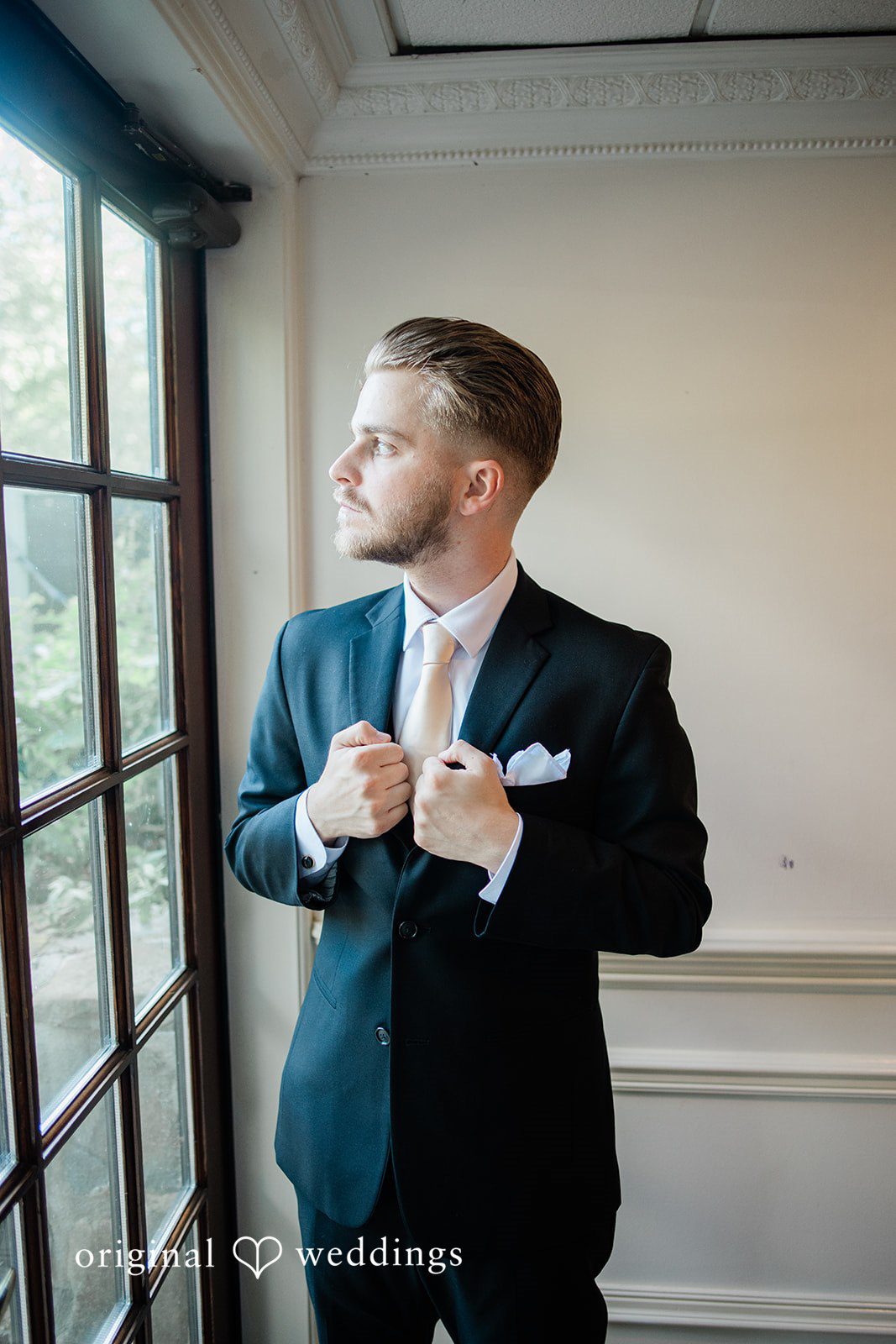 Julia + Camden A groom wearing a black suit and bow tie adjusts his jacket near a window.