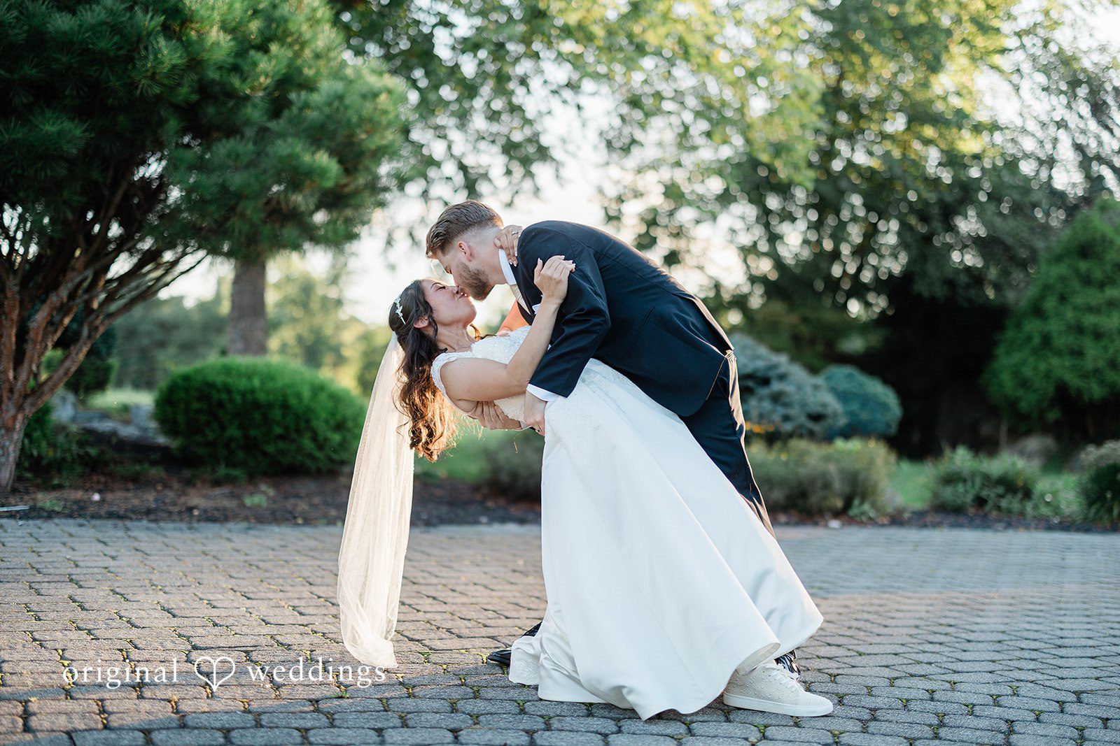 Julia + Camden A groom dips his bride for a kiss on a walkway surrounded by trees.