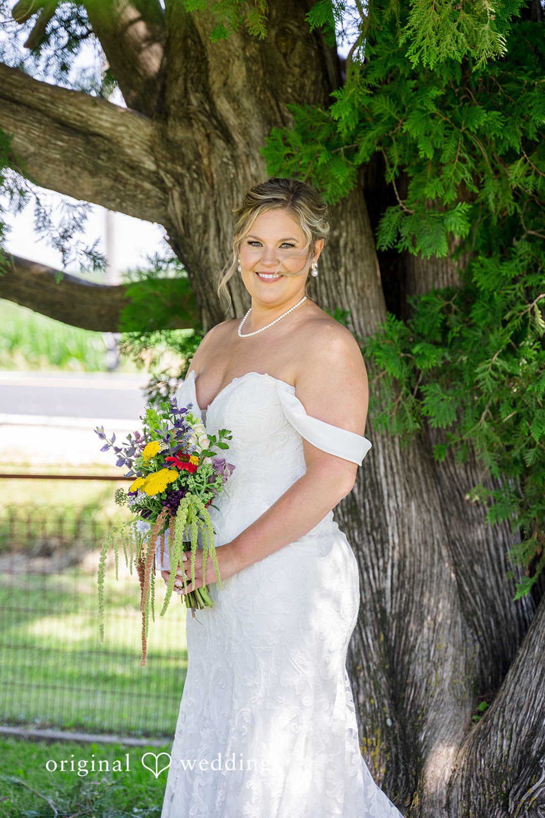 A bride in a white wedding gown smiling warmly while standing in front of lush green trees.