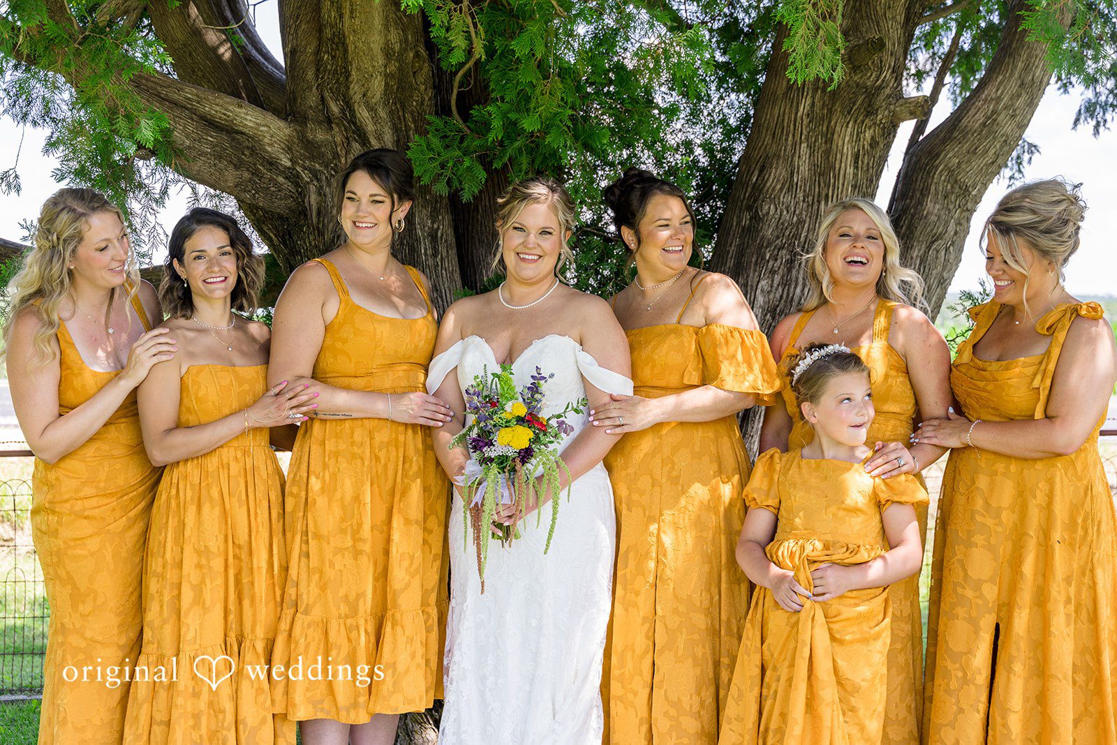 A bride standing together with her bridesmaids wearing matching mustard yellow dresses under a tree canopy.