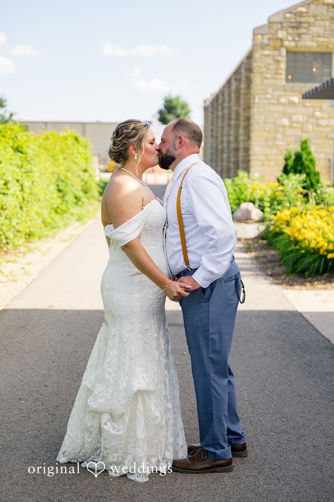 A bride and groom standing hand-in-hand down a paved path near a stone building.