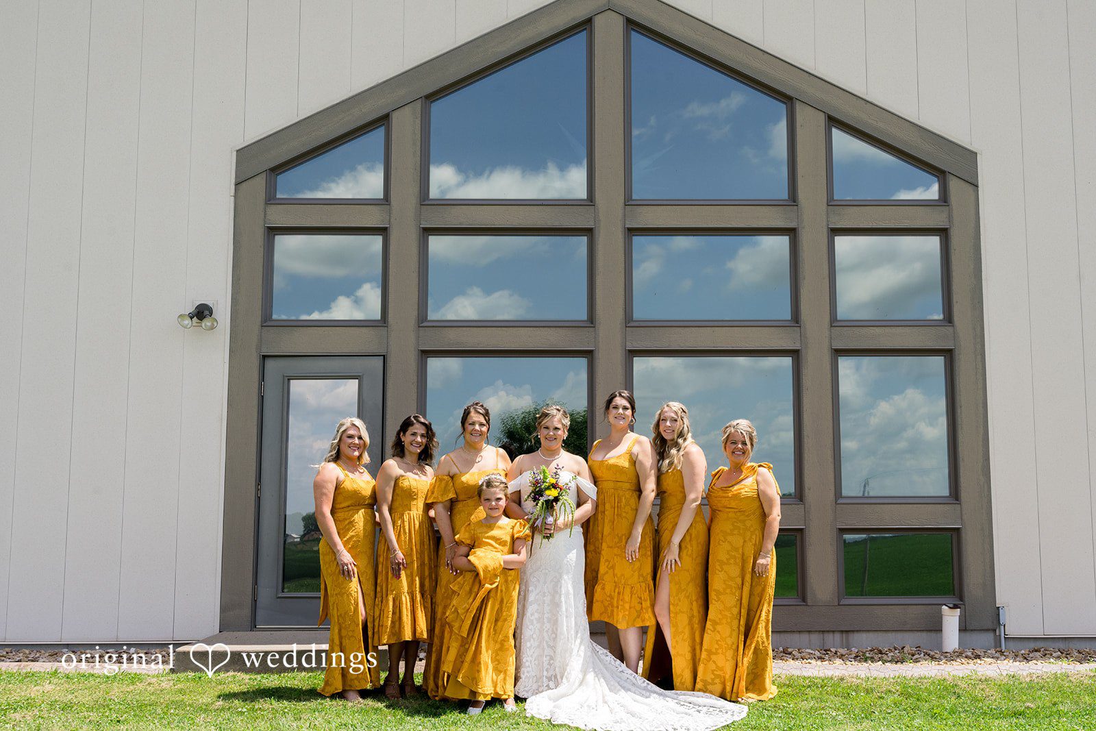A wedding party in mustard yellow attire posing together in front of a modern building with large glass windows.
